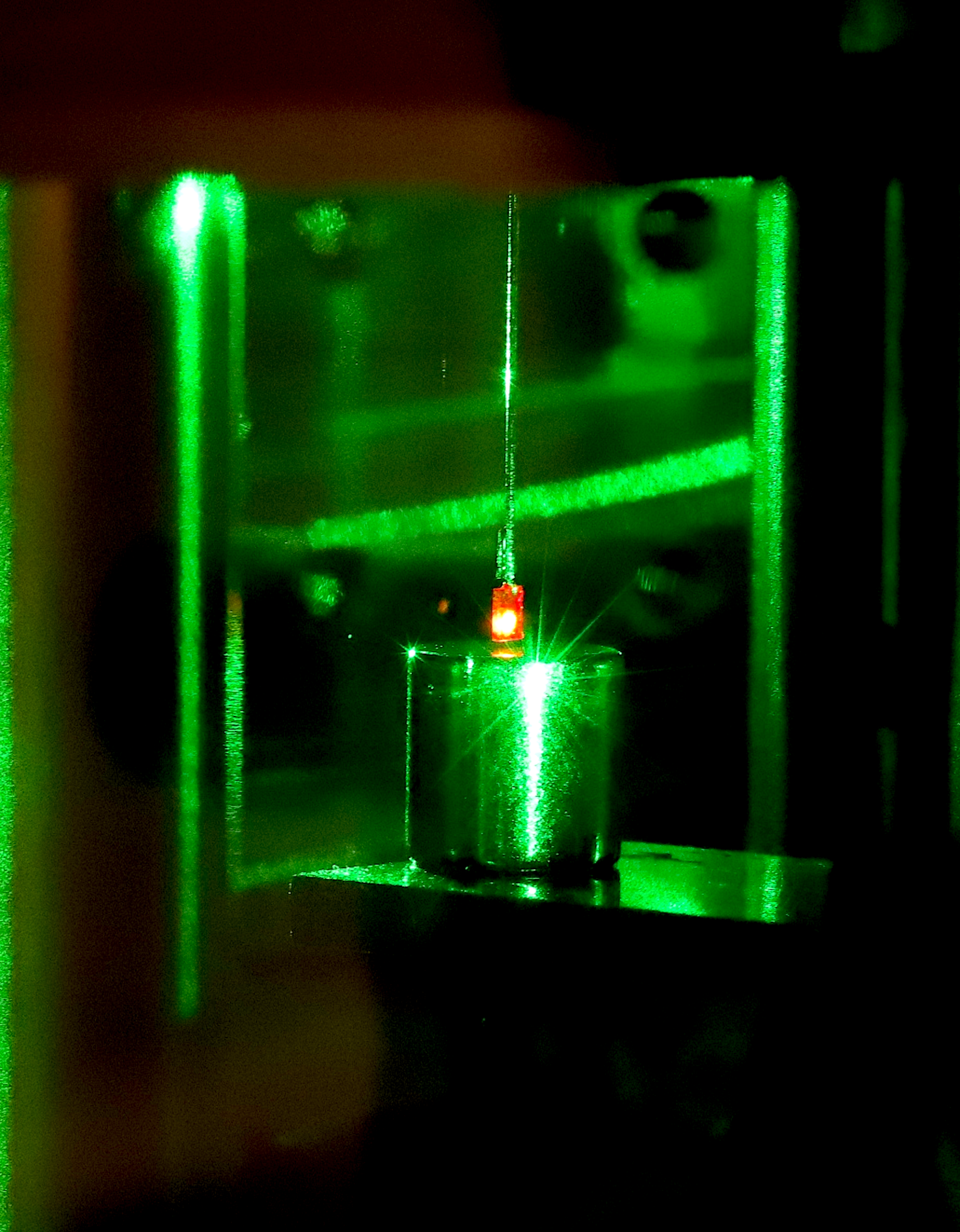 Real-color photo of the lower half of the experimental setup. The diamond with nitrogen-vacancy centers is suspended via a carbon fiber rod above a magnet. When the diamond is illuminated with a green laser, the spin state within the diamond shifts, pushing it down towards the magnet. The diamond glows red due to electrons in the nitrogen-vacancy centers being excited to a higher energy state by the laser.